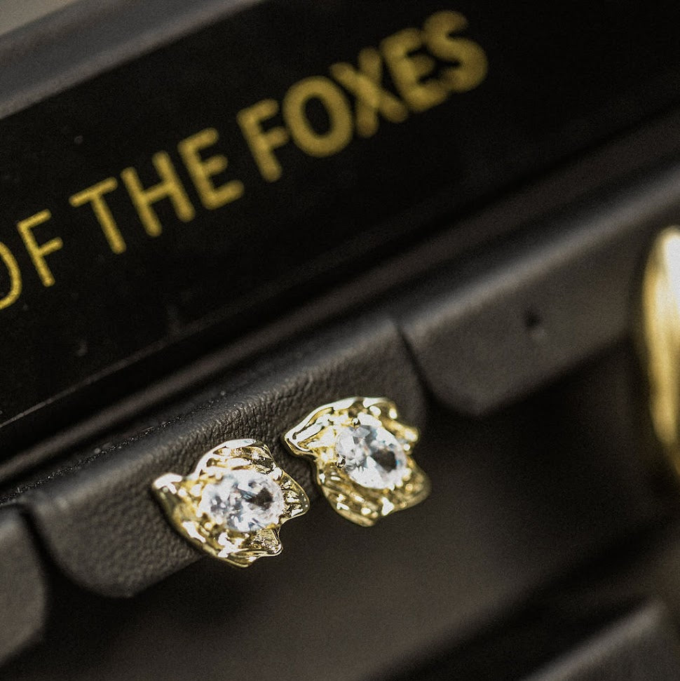 Gold earrings displayed on a black stand with 'Queen of the Foxes' branding.