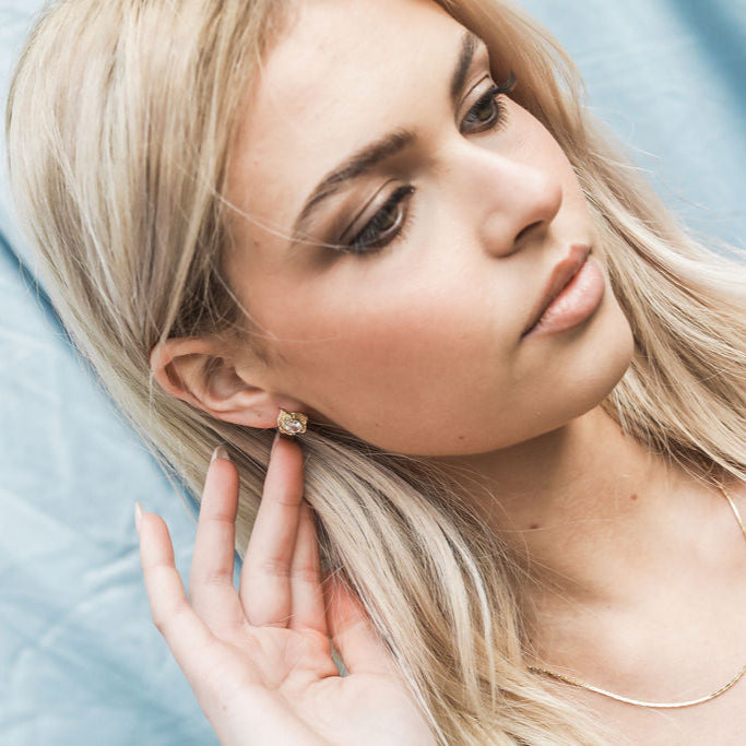 Woman with blonde hair and earrings, wearing a gold necklace, against a light blue background.