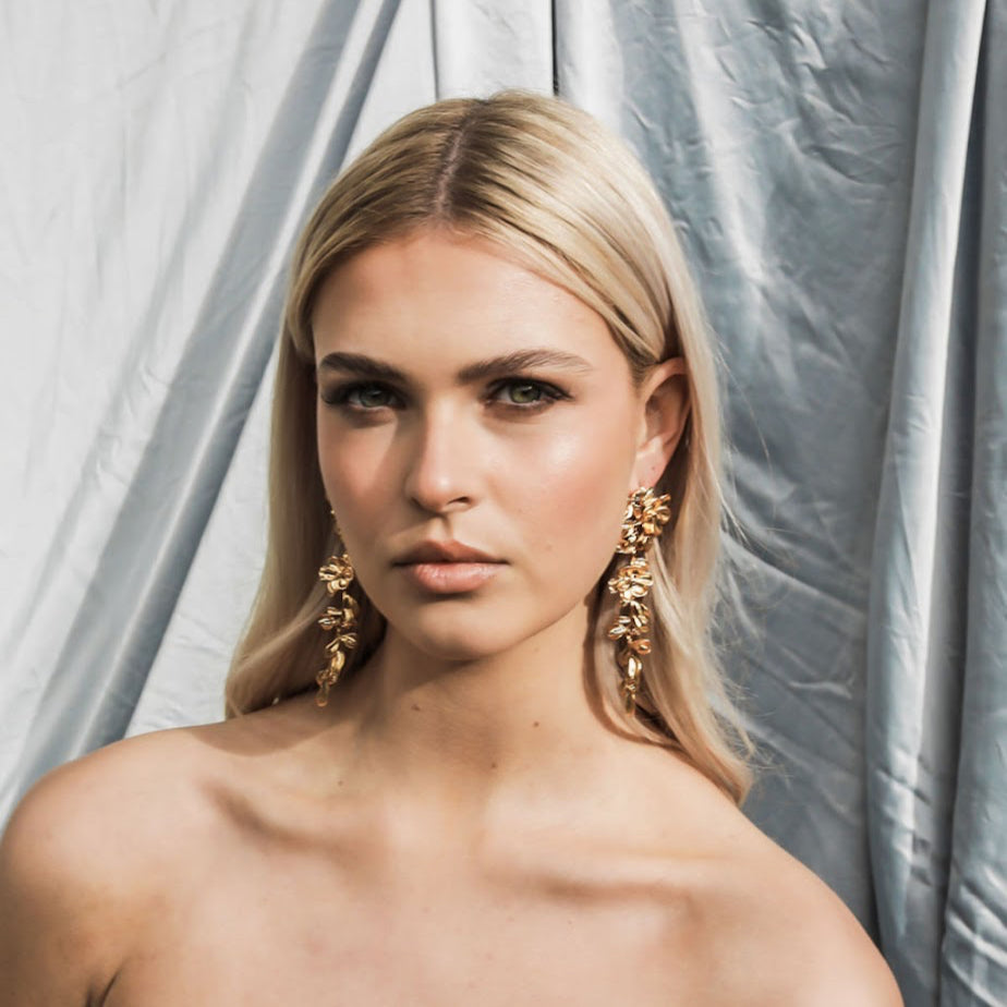 Woman wearing gold floral earrings against a light grey curtain background