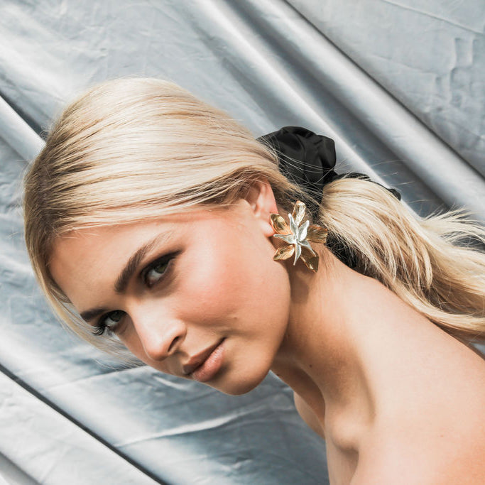 Woman with blonde hair styled in a ponytail with scrunchie and floral statement earrings against a grey draped fabric background
