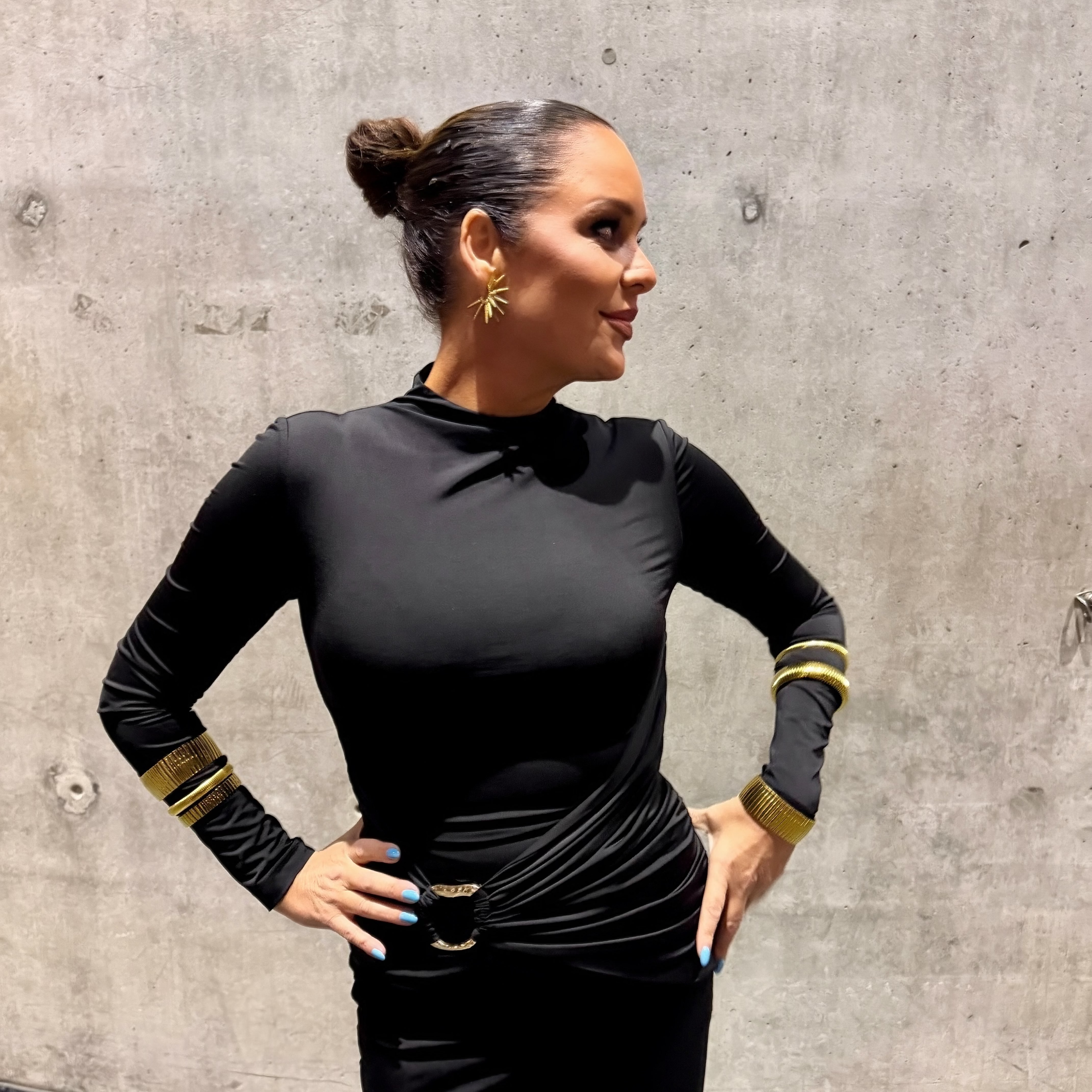 A woman posing in a black long-sleeve mock-neck dress with gold bracelets on the cuffs and a gold belt, standing against a concrete wall.