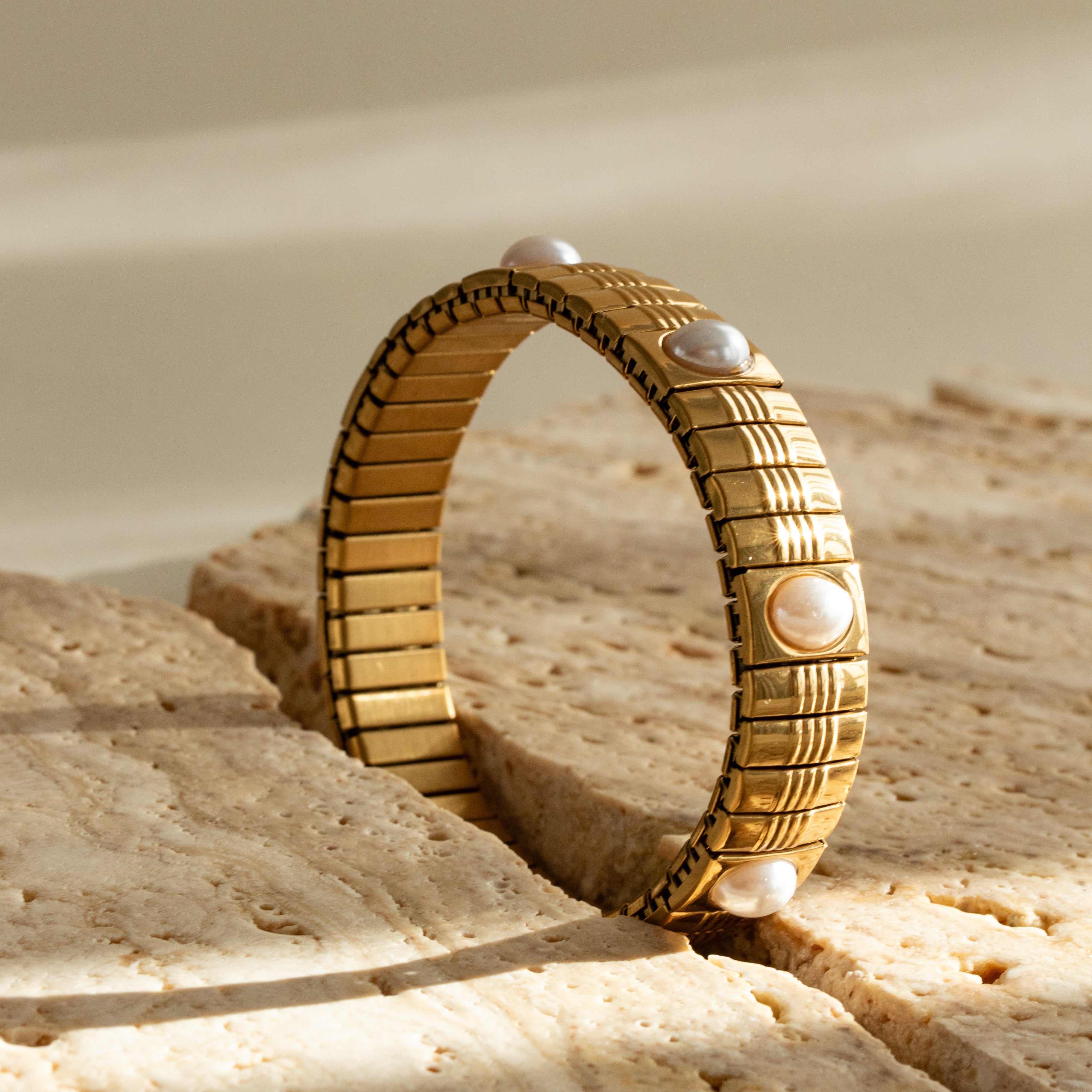 Gold strap and pearl bracelet with textured design on a beige sand-like surface