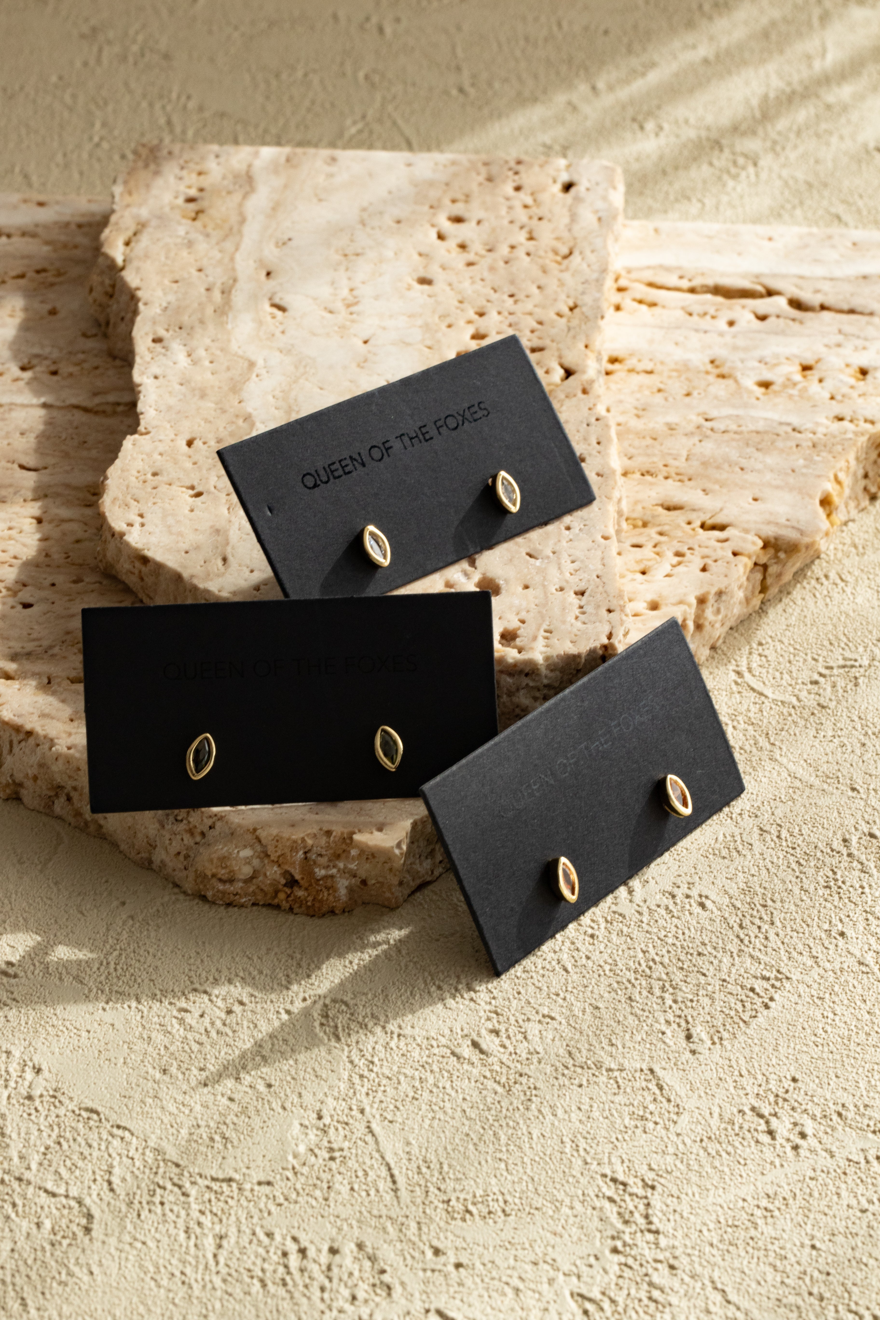 Gold earrings on black card with 'Queen of the Foxes' branding, placed on a textured stone surface.