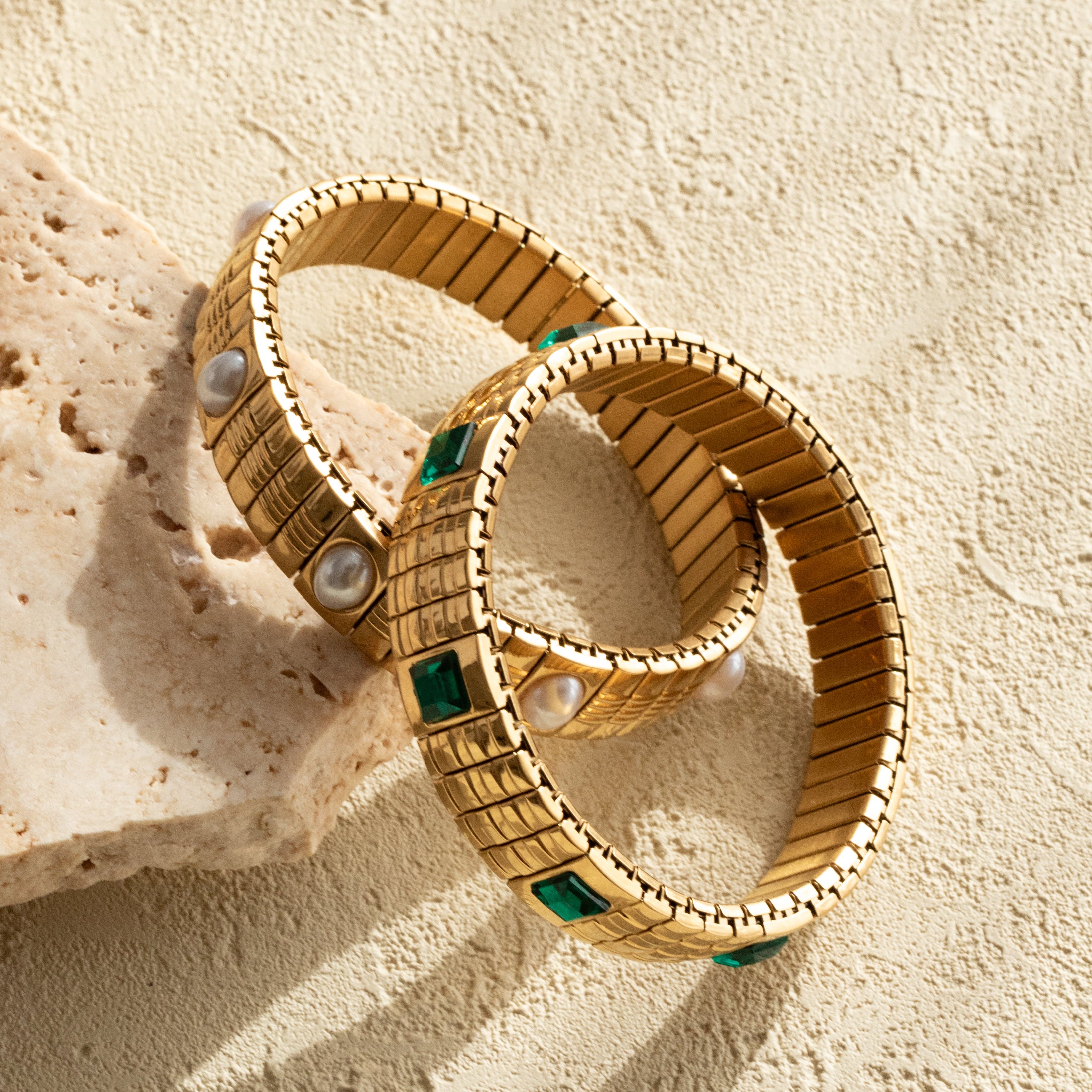 Gold strap bracelets with emerald and pearl accents on a beige stone surface