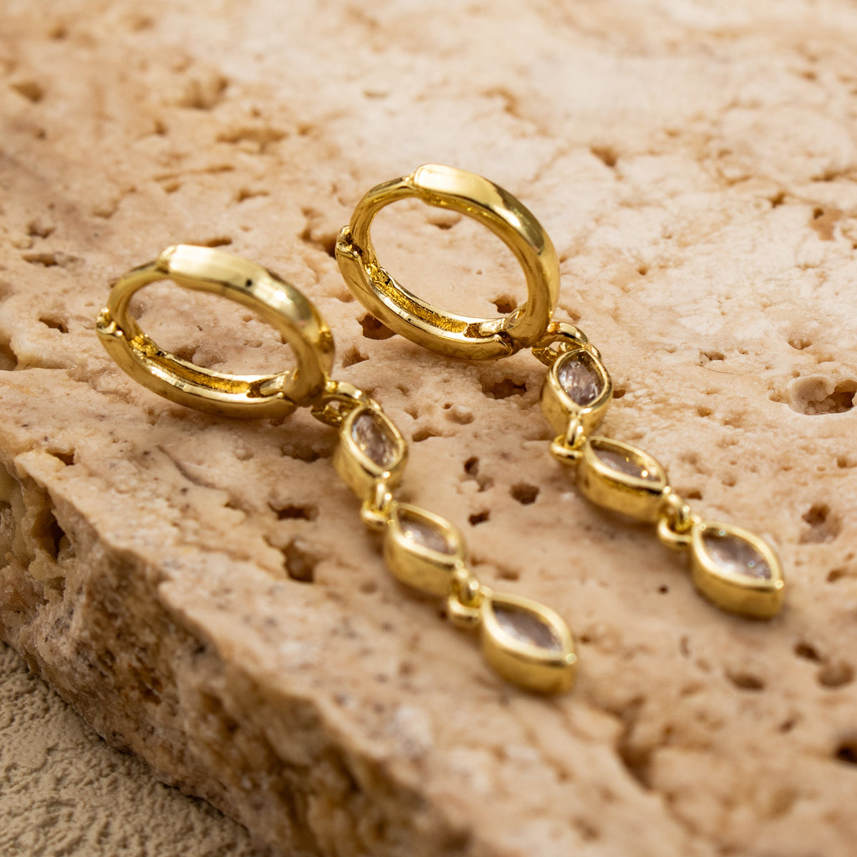 Close-up of Marquise Triple Drop earrings on pale stone, showing hinged gold-plated hoops and linked rub-over stone settings.
