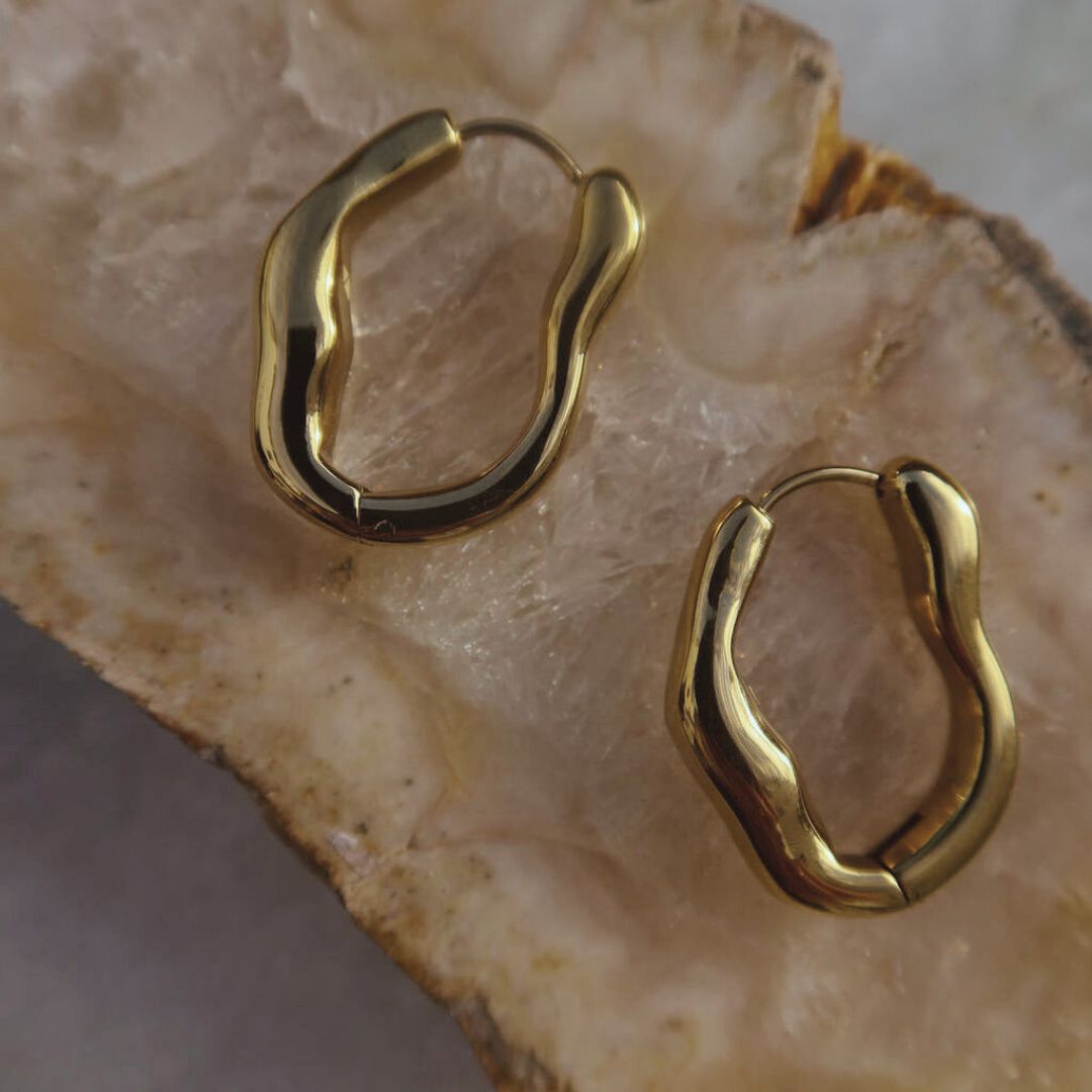 Pair of gold Organic Hoop Earrings on pale stone, showing their irregular oval shape and hinged closures.