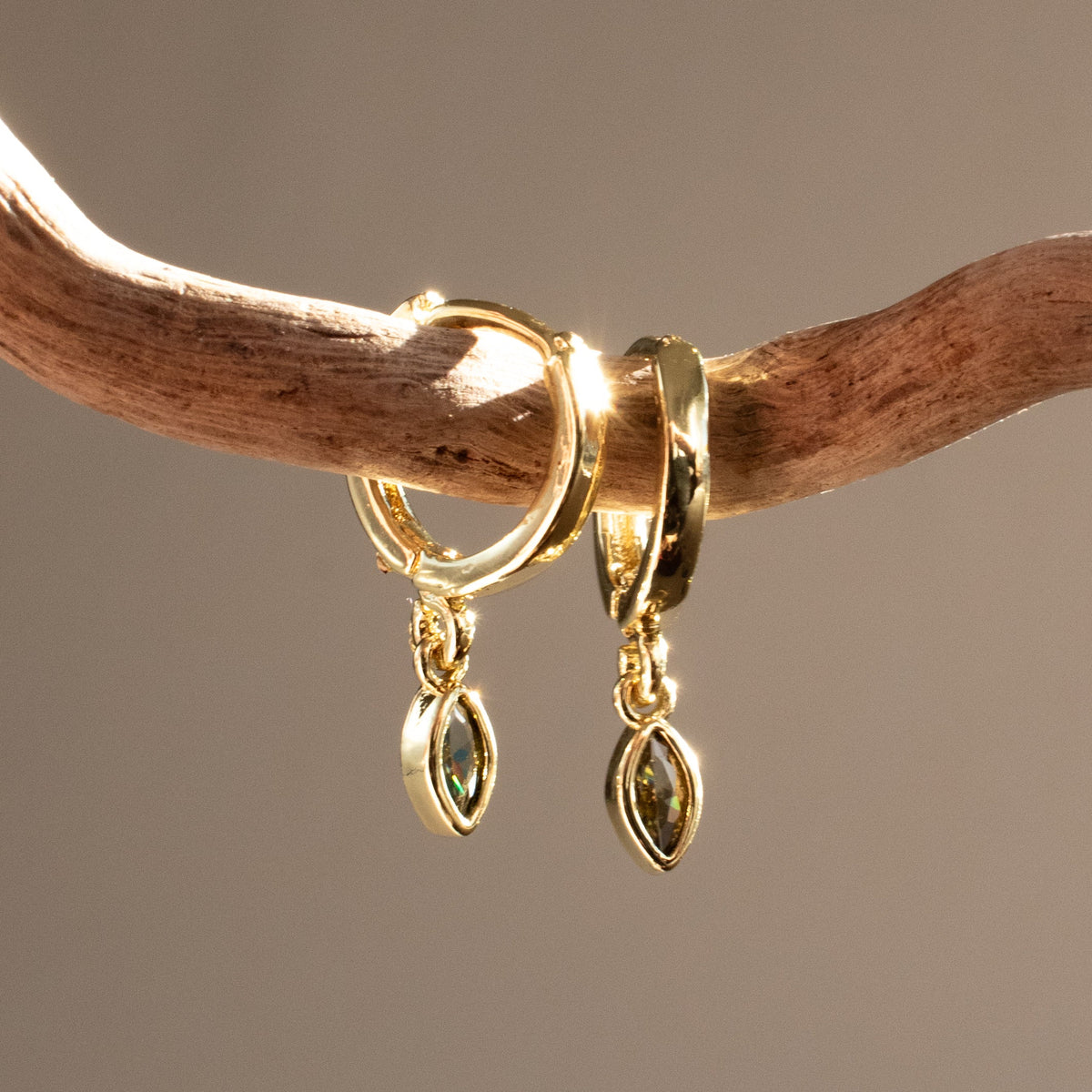 Gold-plated Marquise Single Drop earrings with peridot-green zirconias hanging from driftwood in sunlight.
