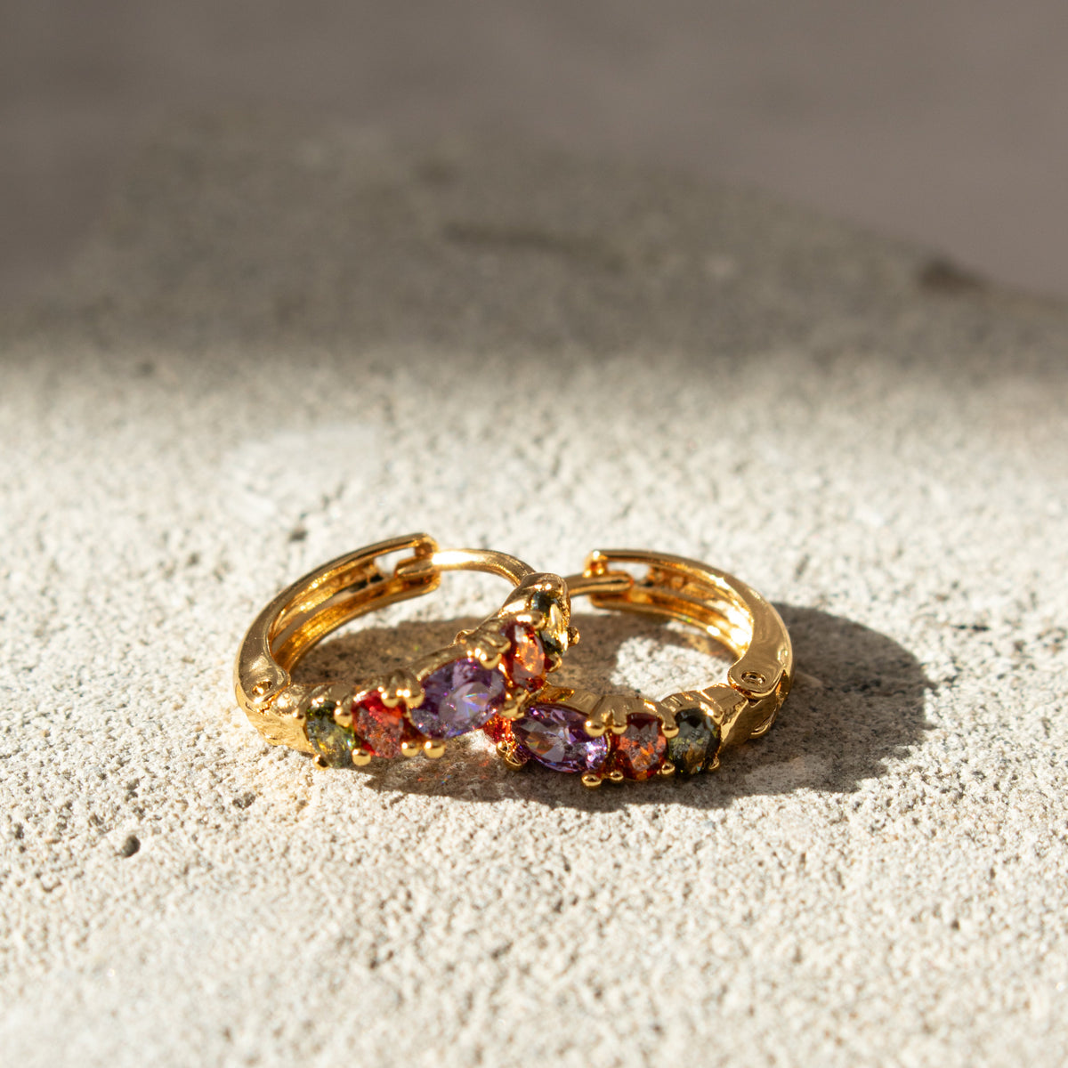 Pair of gold Rainbow Sleeper Earrings resting on pale textured stone in sunlight, with multicoloured zirconias.