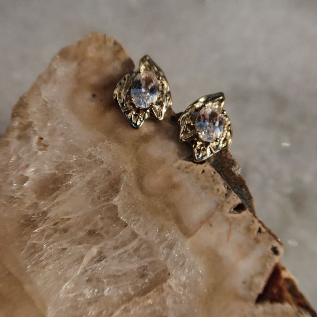 Pair of gold Rough Diamond Stud Earrings resting on a crystal, showing their textured settings and clear oval stones.