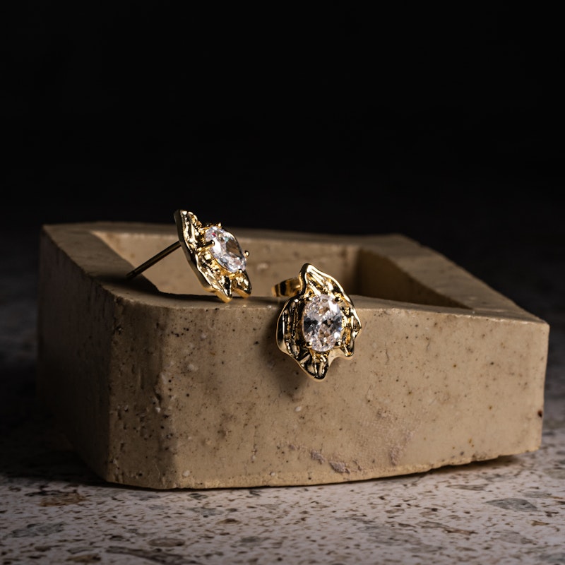 Rough Diamond Stud Earrings on a stone display, featuring clear oval cubic zirconias in irregular gold settings.