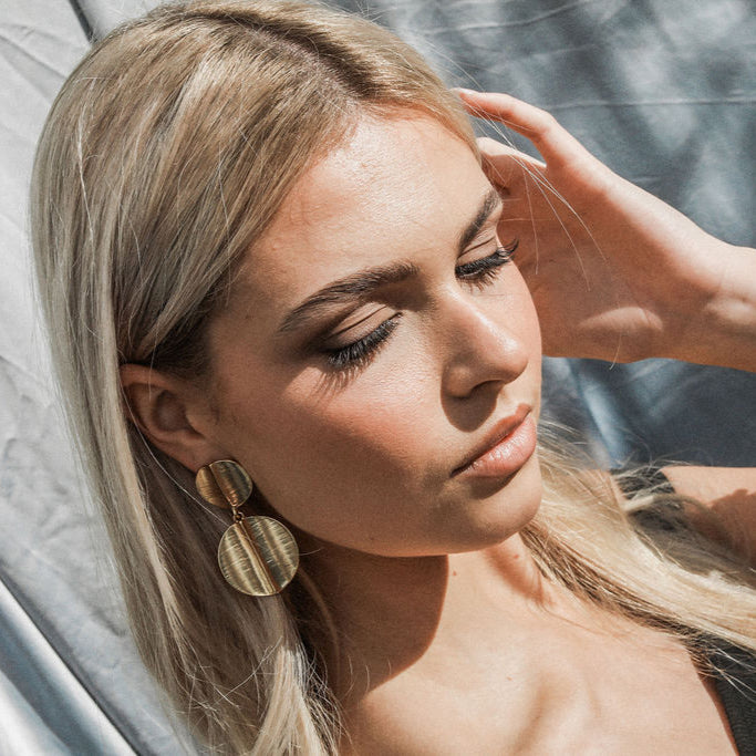 Woman with blonde hair and gold earrings, wearing a black top, sitting on a white surface.