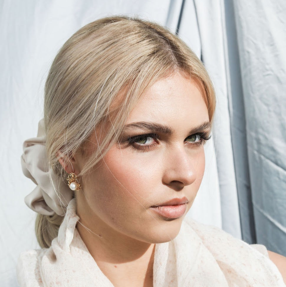 Woman with styled hair and soft pink scarf, scrunchie and pearl earrings  against a neutral background