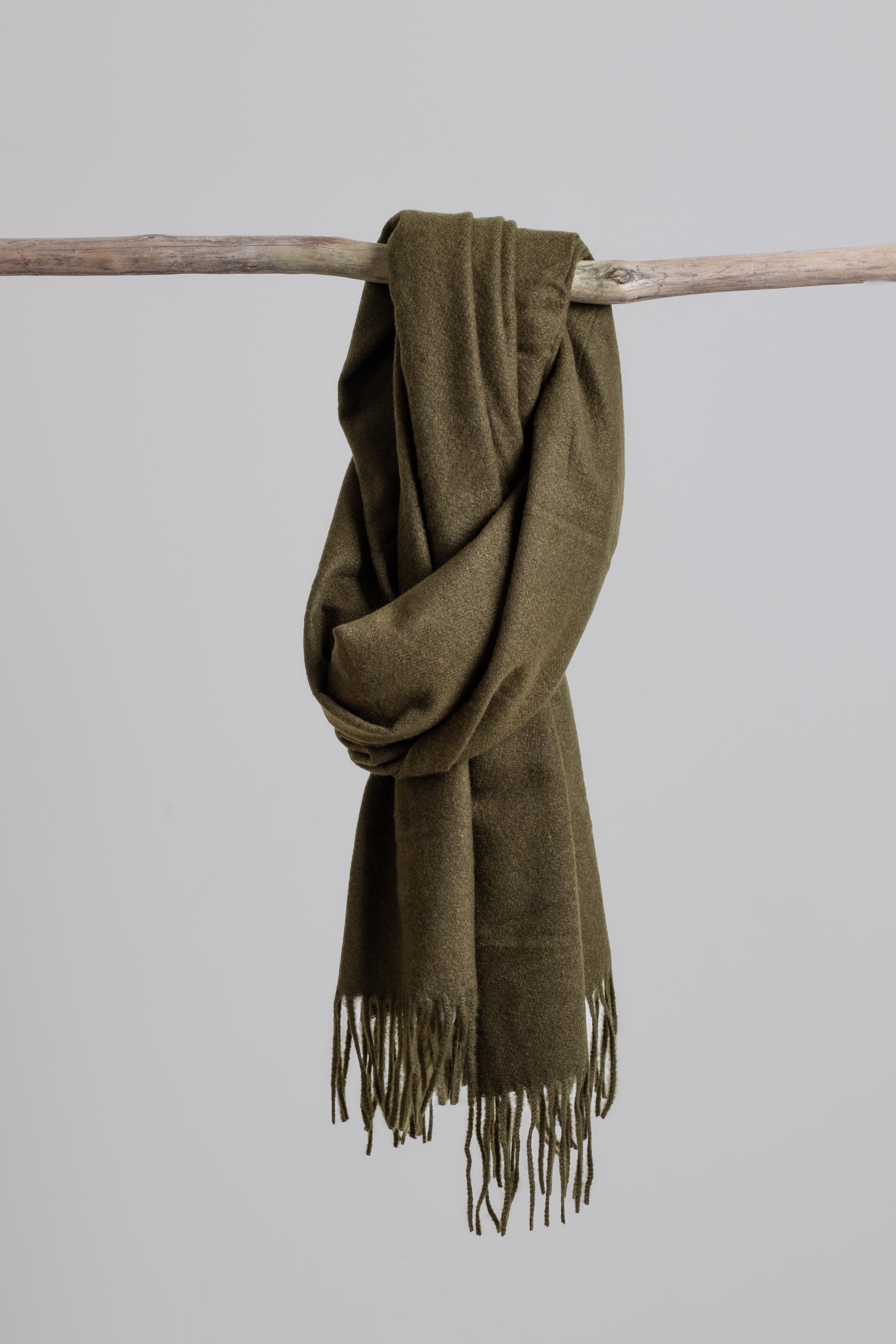 Olive green scarf with fringes draped over a branch against a light gray background