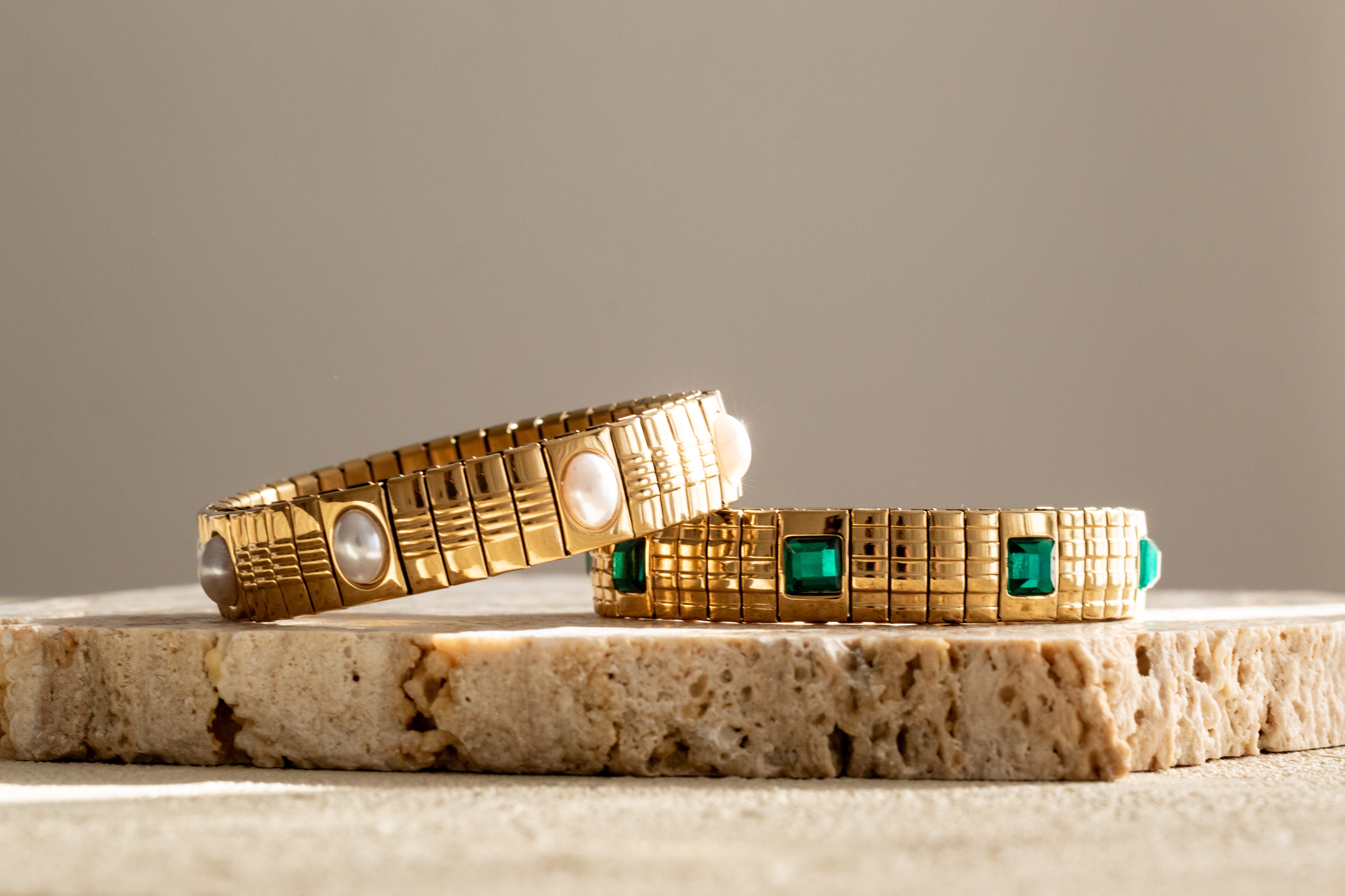 Gold bracelets with emerald and pearl gemstones on a textured surface