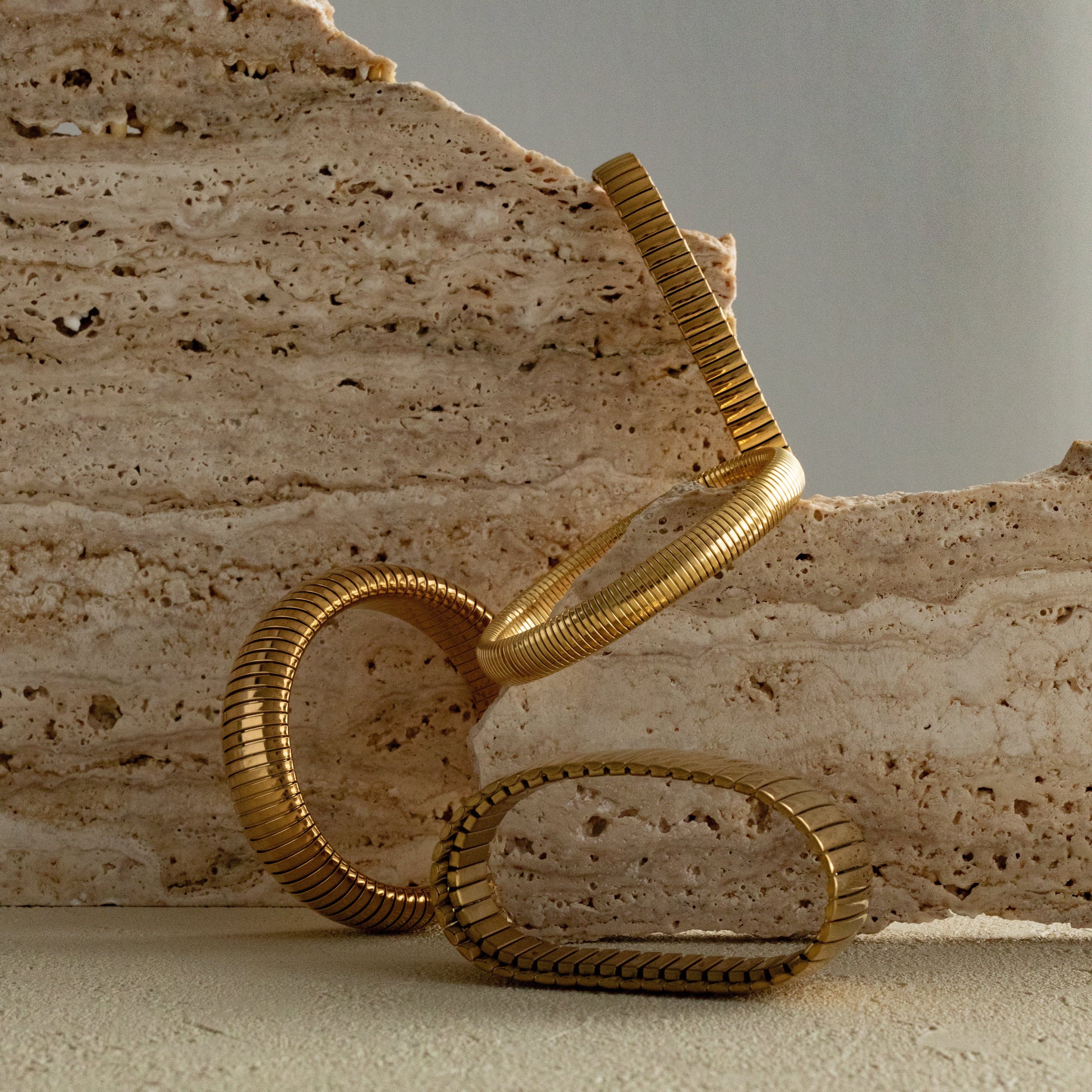 Gold flexi and strap bracelets sculpture on a textured beige surface