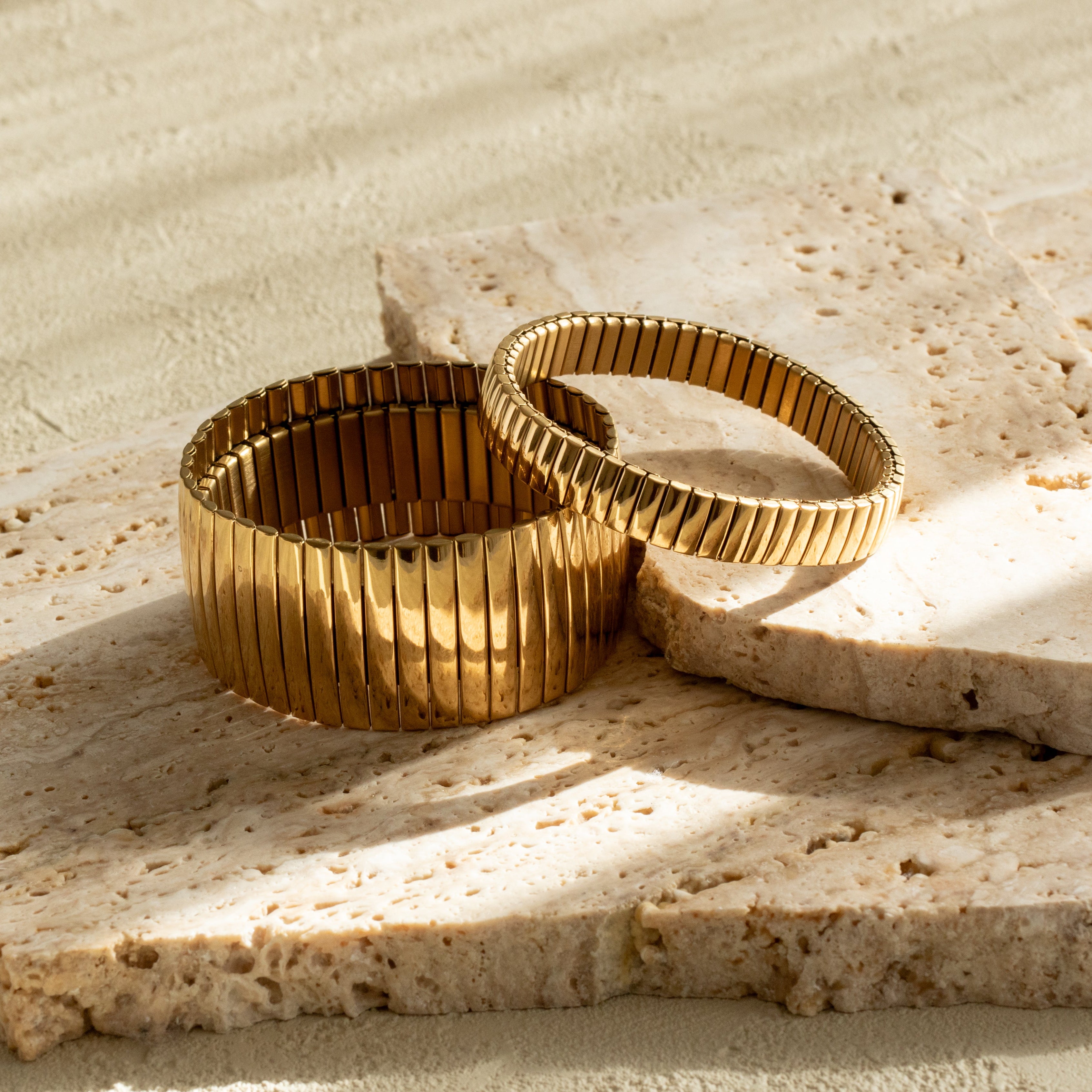 Two gold flexible strap bracelets on a textured stone surface with sand background