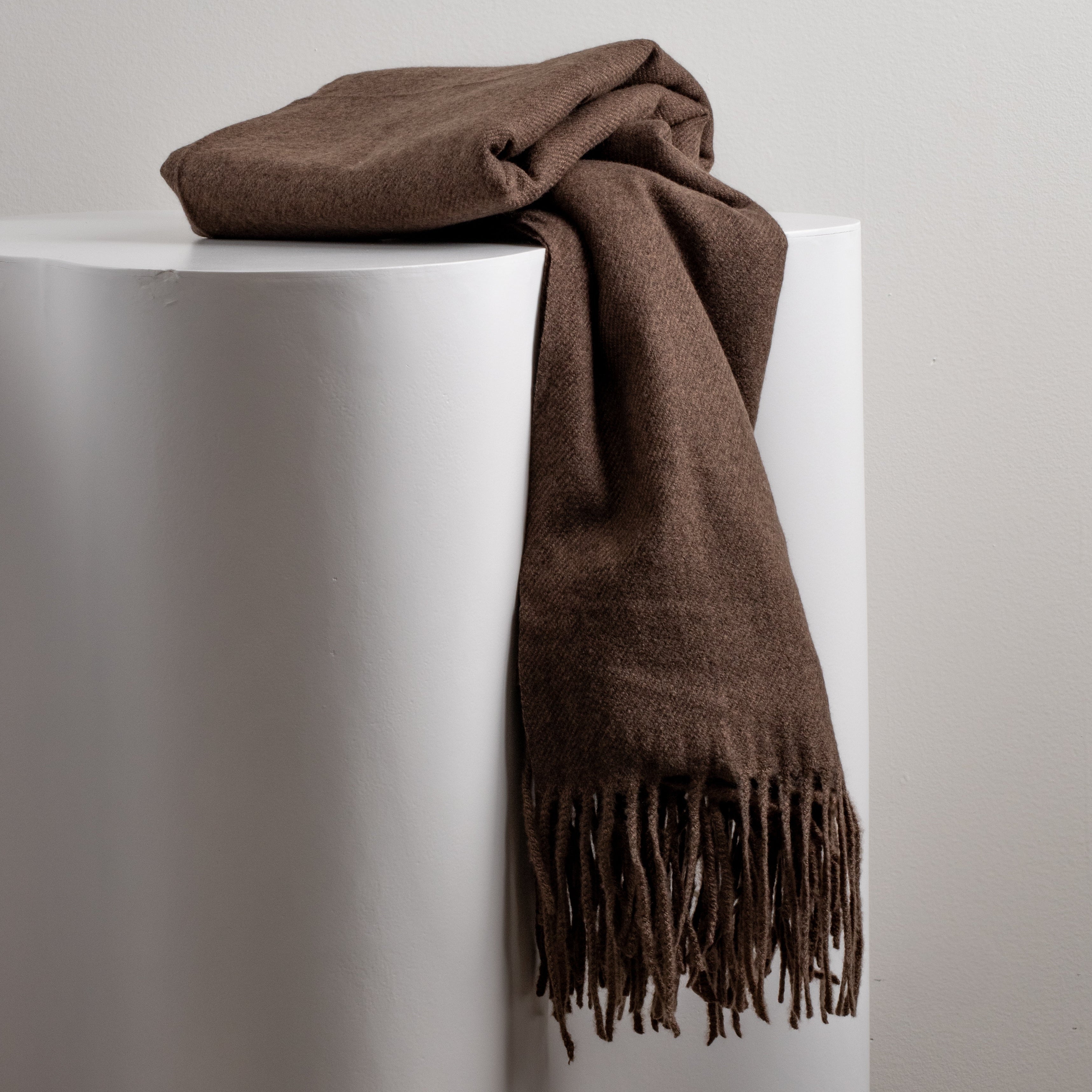 Chocolate brown scarf with fringe on a white pedestal against a light gray background