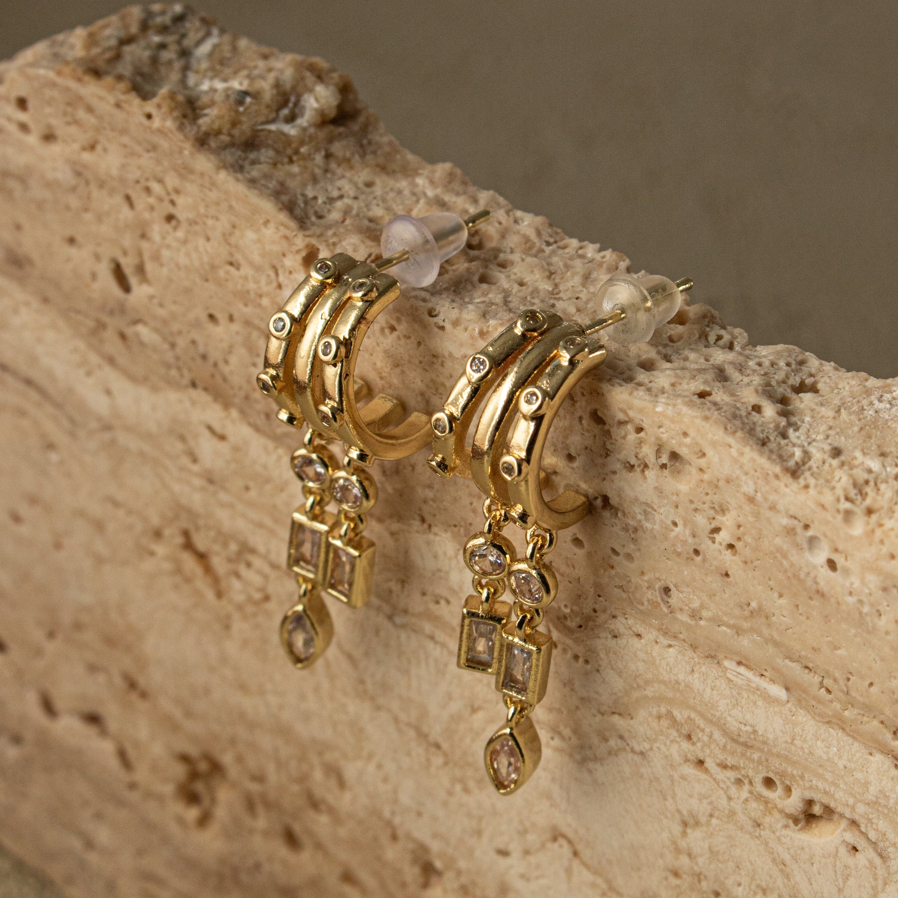 Gold hoop earrings with dangling elements on a textured beige surface