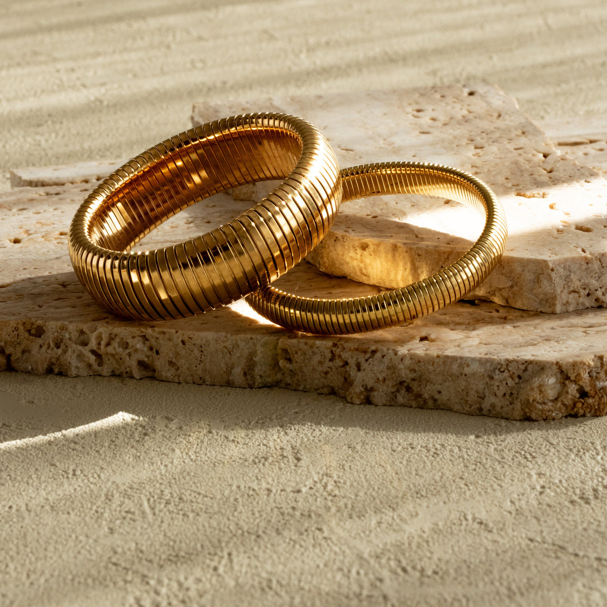 Thick and medium Queen of the Foxes gold-plated banded bracelets displayed together on pale stone, showing their different widths and ribbed finish.