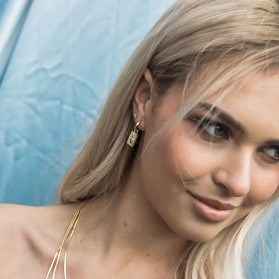 Blonde model wearing a gold-plated Dewdrop Sleeper Earring with layered necklaces against a blue background.