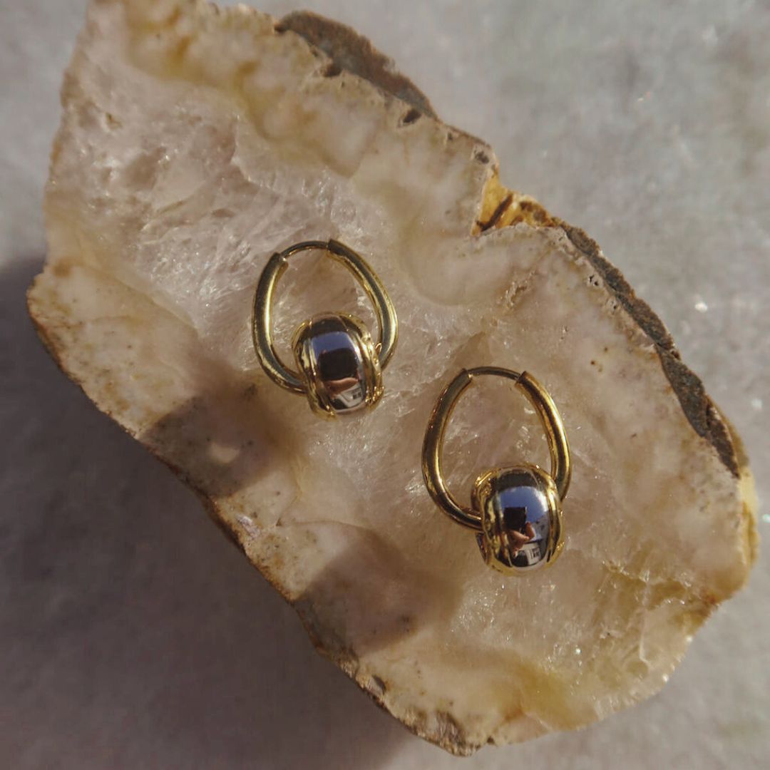 Double Barrel Love earrings with gold hoops and polished silver-tone barrel charms edged in gold, displayed on natural stone.