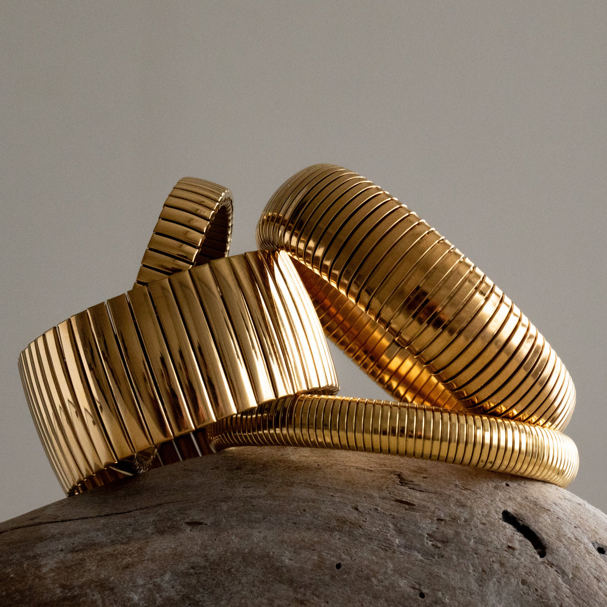 Thick and medium Queen of the Foxes gold-plated banded and flexi strap bracelets displayed together on pale stone, showing their different widths and ribbed finish.
