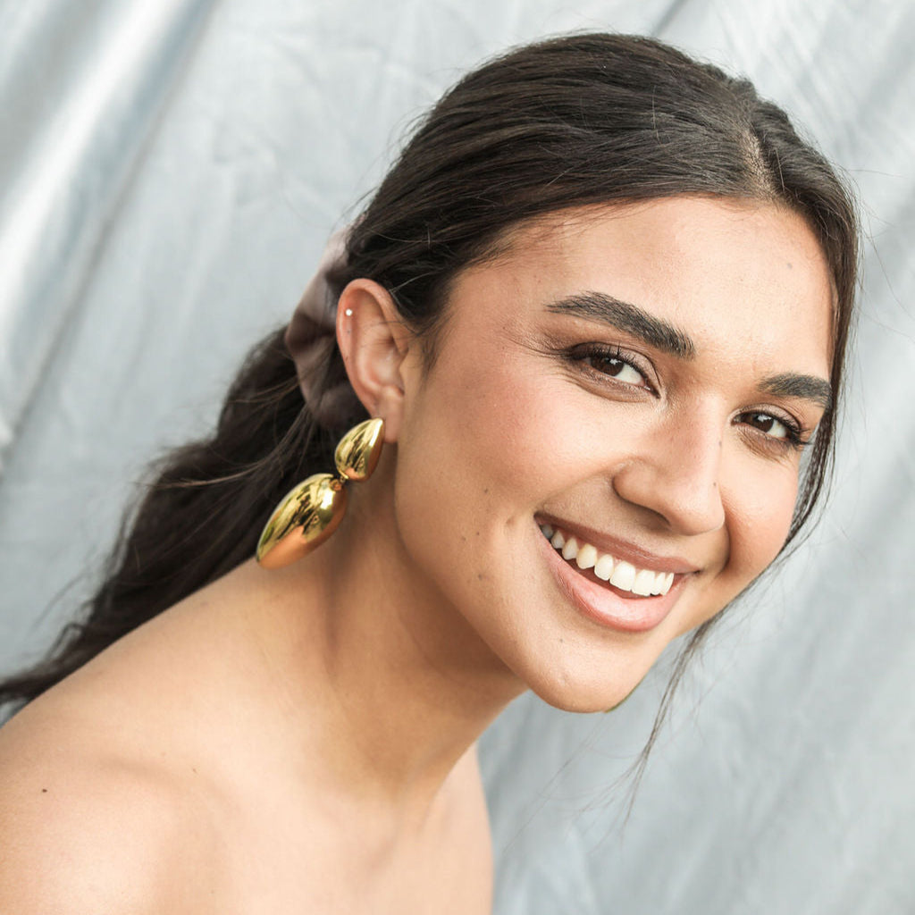 Smiling brunette wearing gold Origin drop earrings, showing their polished two-part shape against a grey backdrop.
