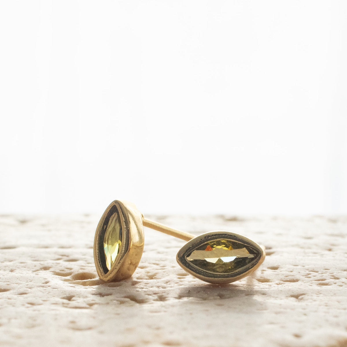 Two Peridot Marquise Stud earrings resting upright and sideways on pale textured stone, with olive-green faceted zirconias.