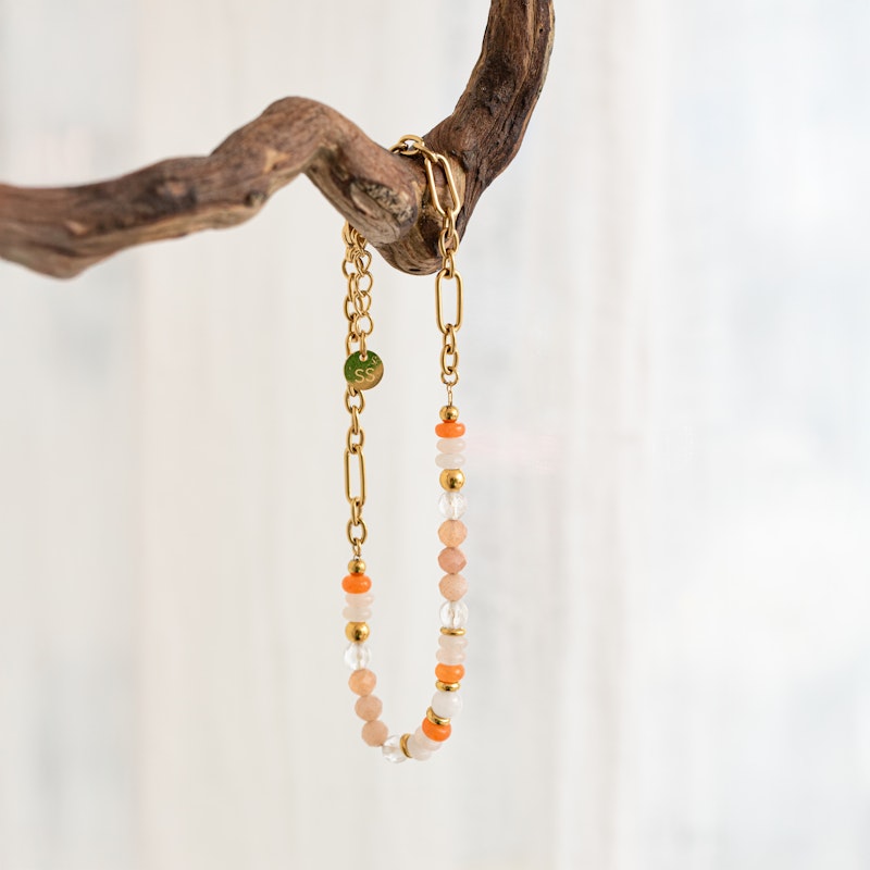 Precious Stones + Chain Bracelet hanging from a branch, showing pastel beads, orange accents and elongated gold links.