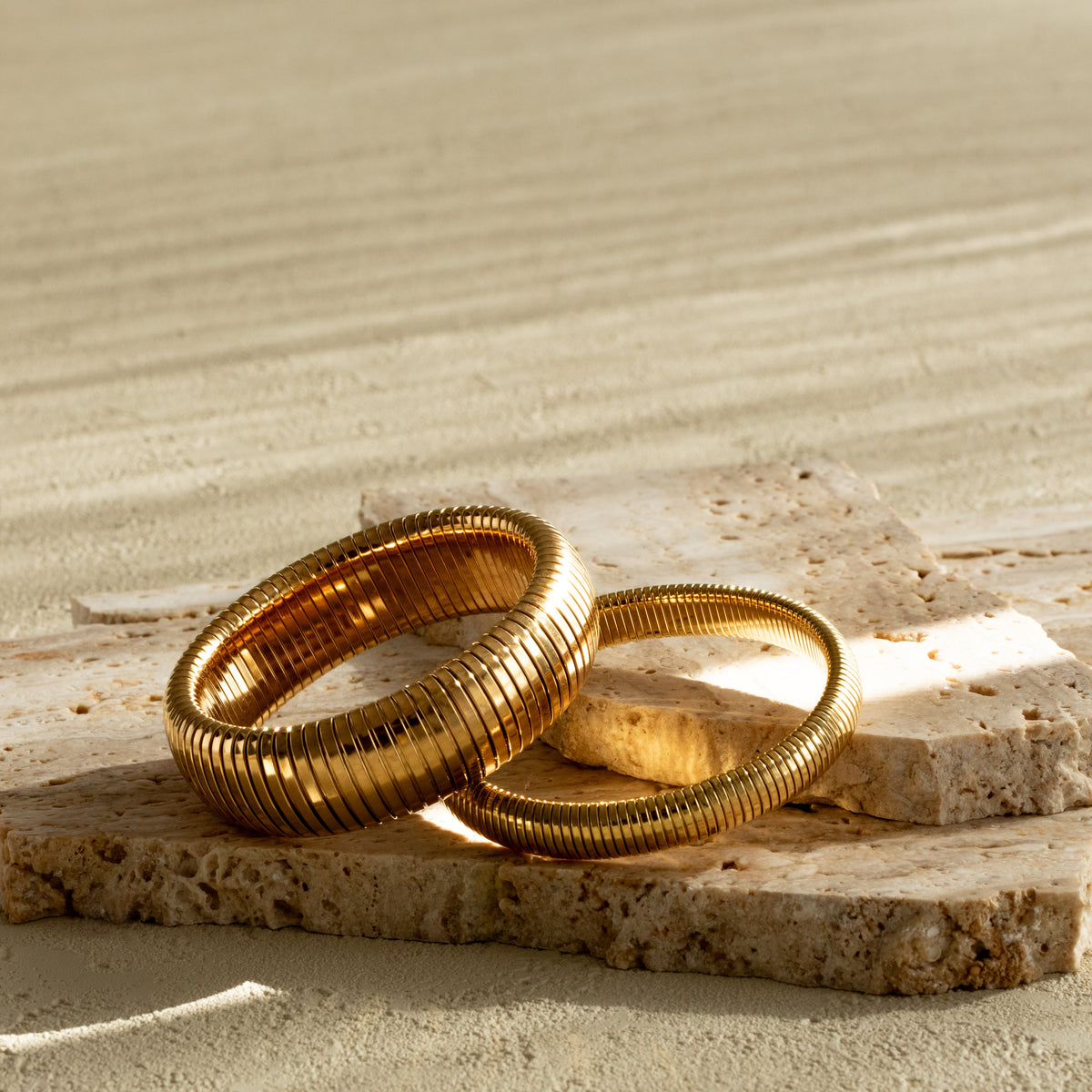Thick and medium Queen of the Foxes gold-plated banded bracelets displayed together on pale stone, showing their different widths and ribbed finish.